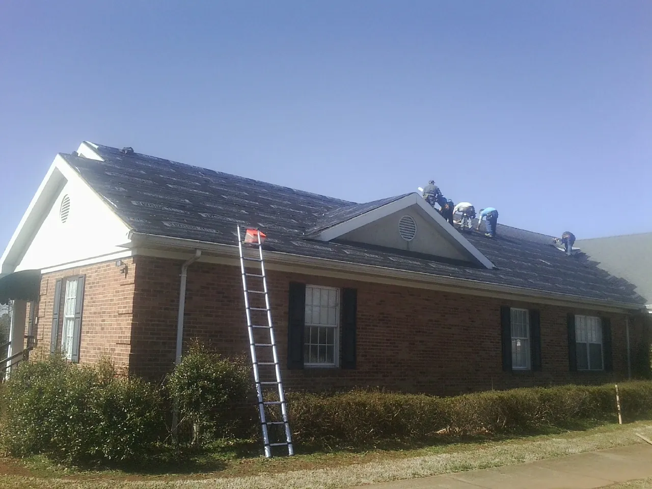 Crew tearing shingles off a brick ranch roof with a ladder set — Metal Roof Repair work by our crew in Centerview