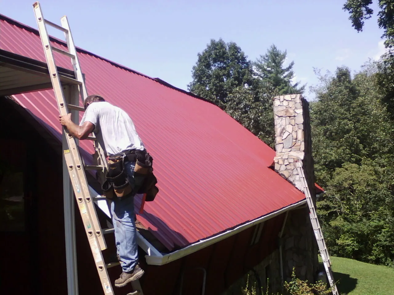 Roofer on a ladder beside a red metal roof and stone chimney — Soffit & Fascia Repair by Roofing Brothers in Centerview
